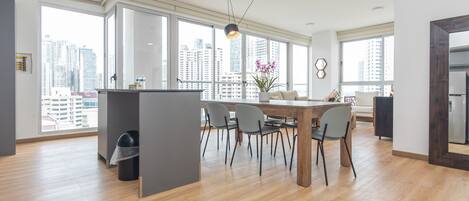 A spacious living area with a dining table, chairs, and a large window offering a city view.