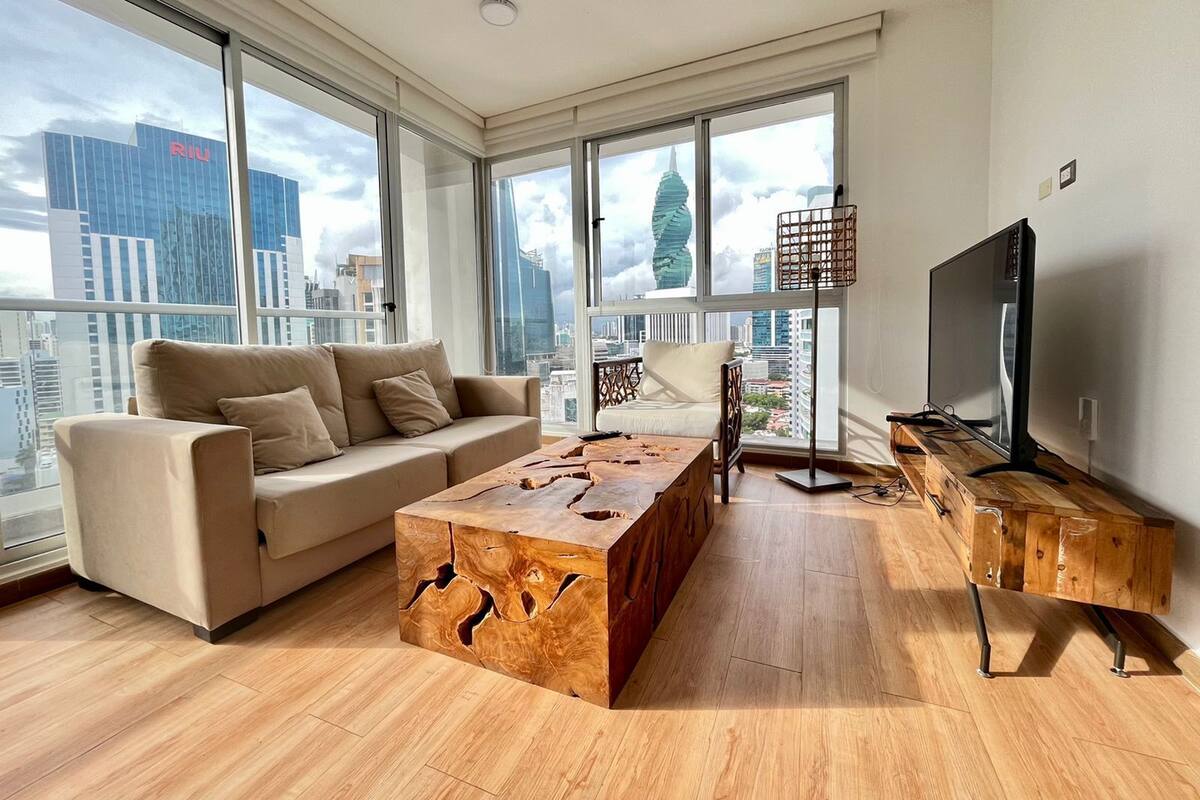 A modern living room with a sofa, coffee table, TV, and large windows offering city views.
