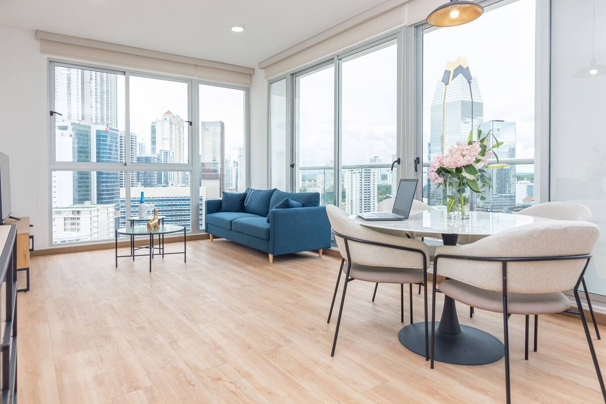 A spacious living room with a large window, a blue sofa, a dining table with chairs, and a laptop on the table.