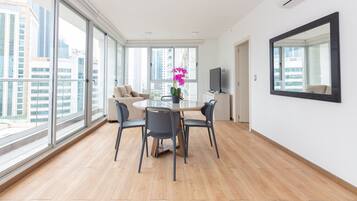 Un salon moderne avec une table ronde en verre, quatre chaises, un canapé, un téléviseur et une grande fenêtre donnant sur la ville.