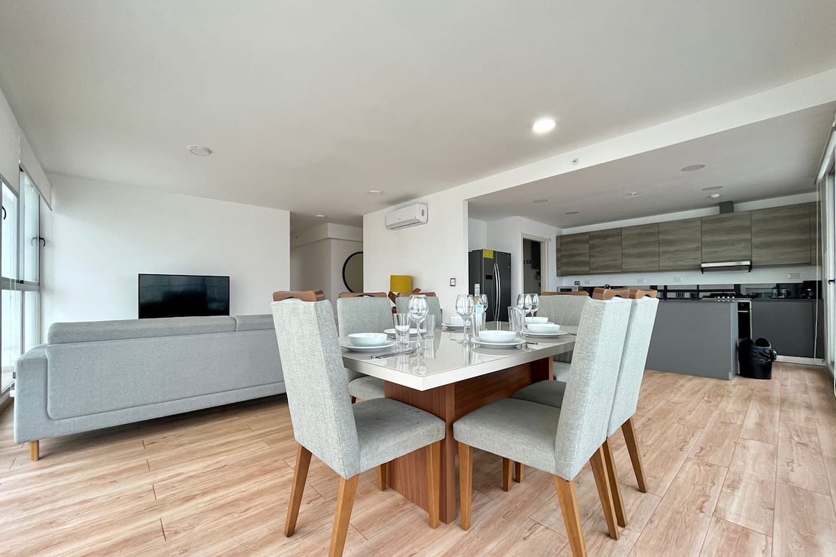 A modern dining area with a wooden table and grey upholstered chairs, a grey sofa, and a kitchen in the background.
