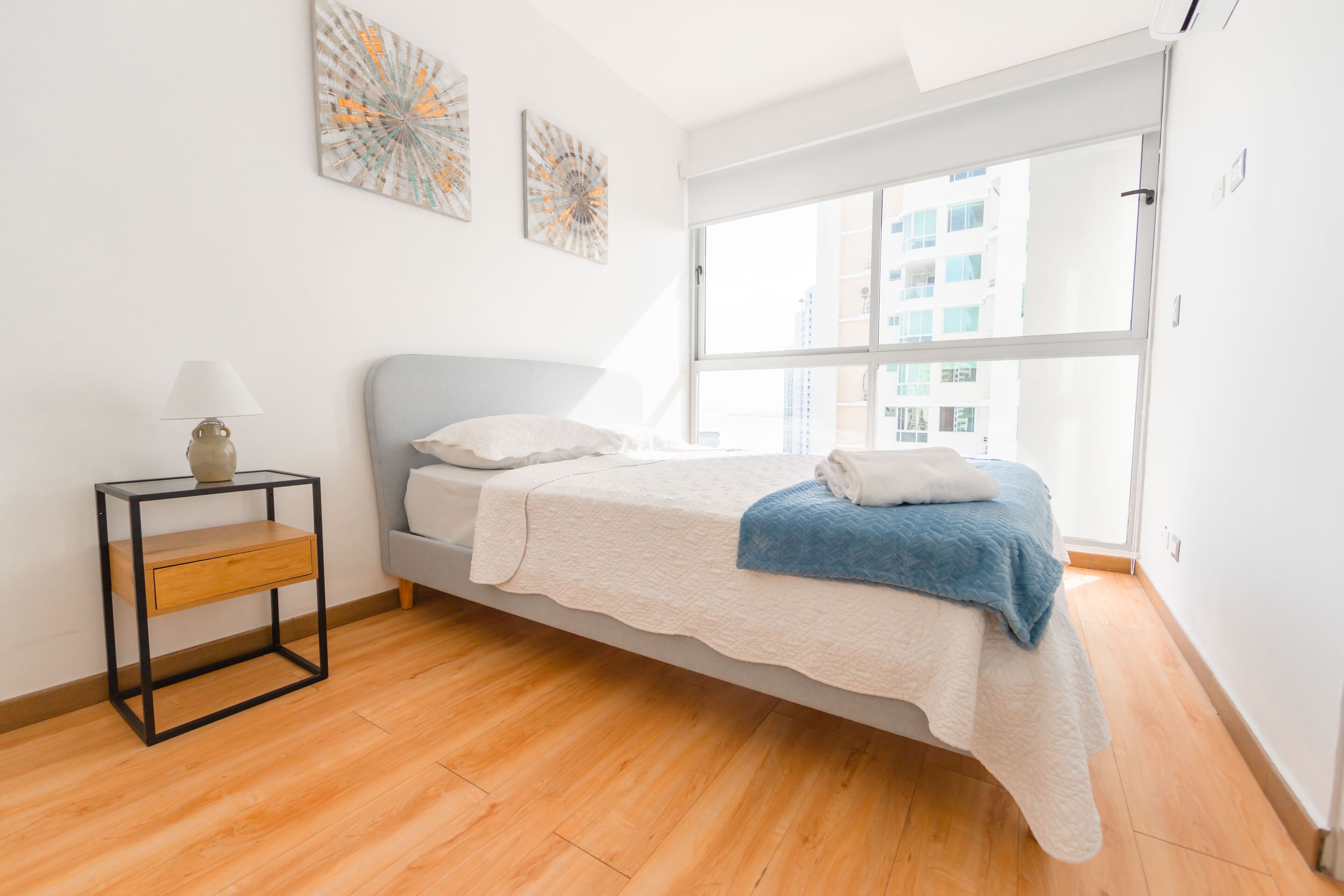 Une chambre à coucher avec un lit, une table de chevet, une fenêtre et des œuvres d’art accrochées au mur.