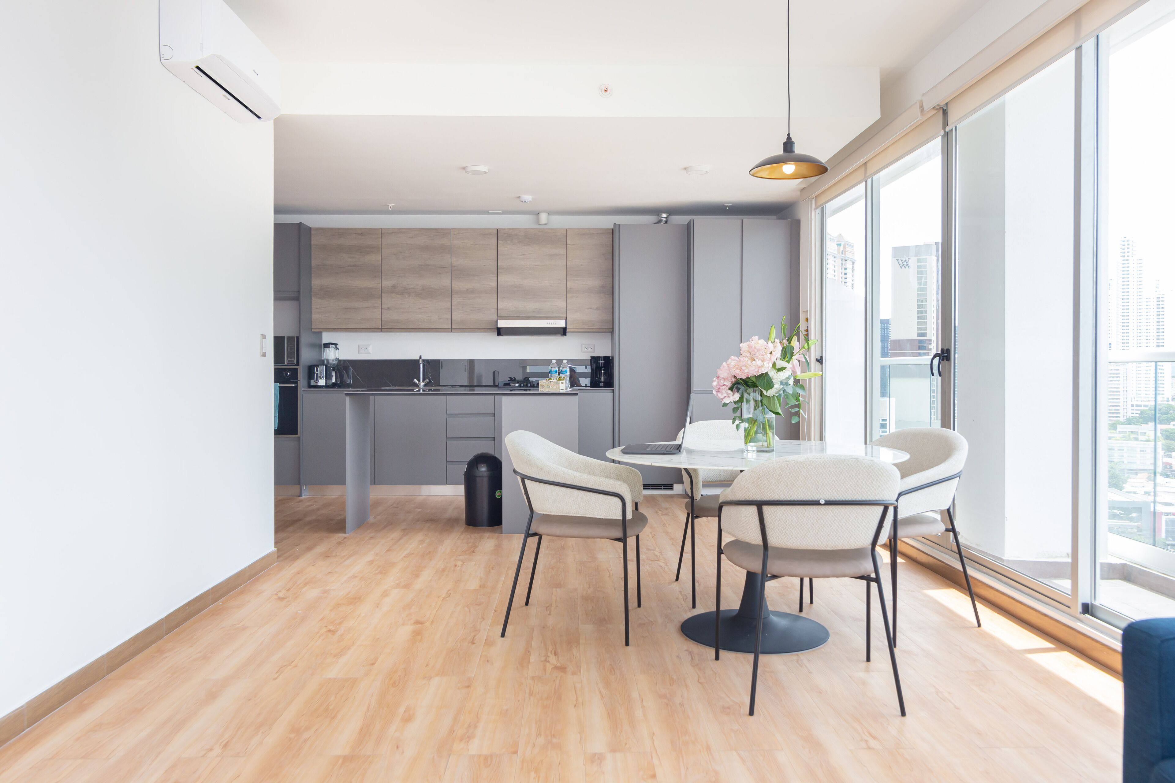 Una cocina moderna con armarios grises, un comedor con una mesa redonda y cuatro sillas, y un ventanal con vistas a la ciudad.
