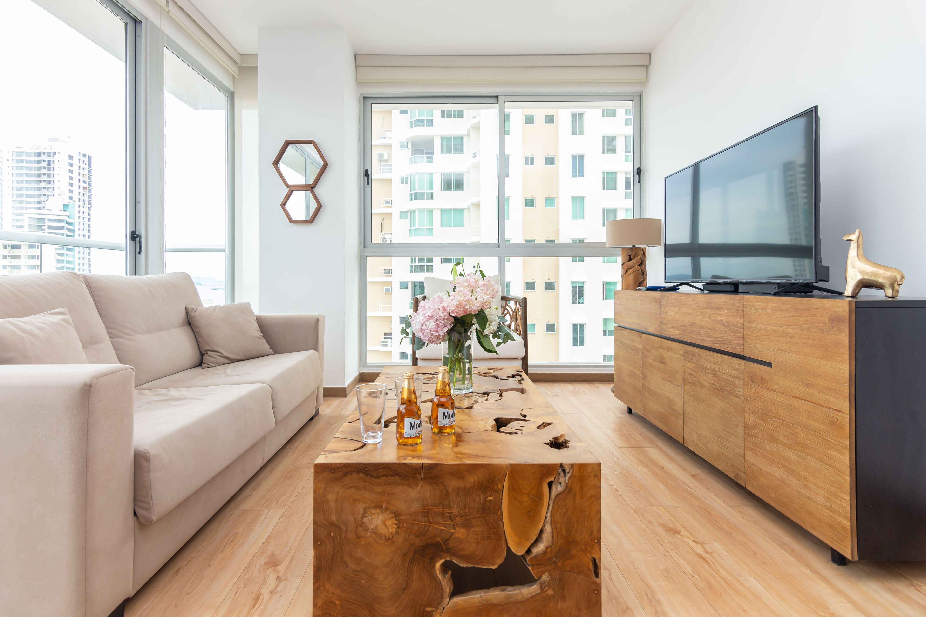 Ein modernes Wohnzimmer mit einer Couch, einem Couchtisch mit Getränken, einem Fernseher und Blick auf Gebäude durch große Fenster.