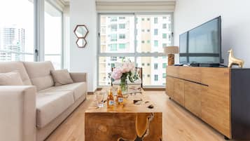 Ein modernes Wohnzimmer mit einer Couch, einem Couchtisch mit Getränken, einem Fernseher und Blick auf Gebäude durch große Fenster.