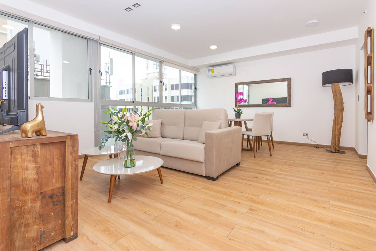 A modern living room with a beige sofa, a wooden coffee table, and a dining area with a white table and chairs.