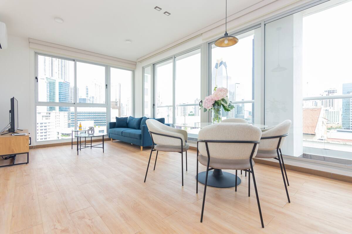 A spacious living room with large windows, a blue sofa, two chairs, a small table, and a TV on a wooden stand.