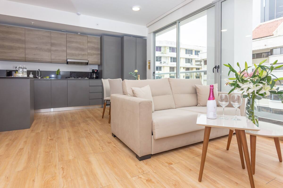 A modern kitchen with grey cabinets, a beige sofa, and a dining table with wine glasses and a vase of flowers.