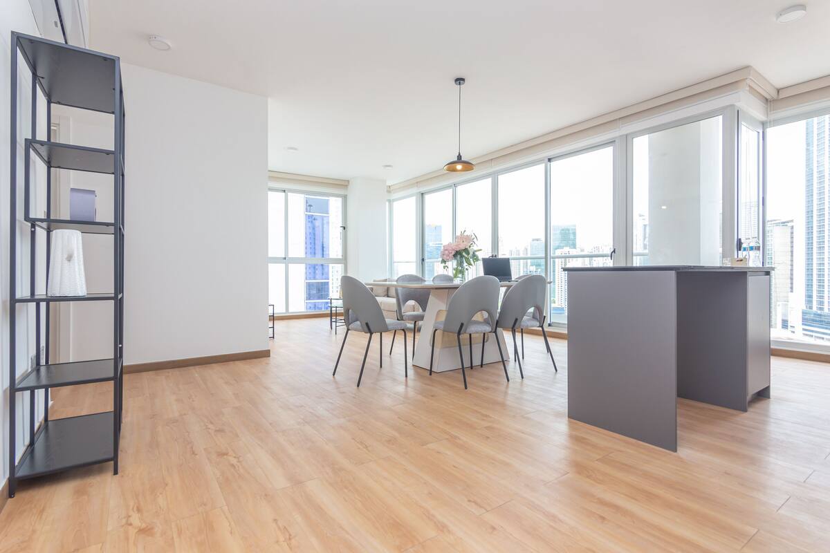 A spacious room with a wooden floor, a dining table with chairs, a bookshelf, and large windows offering a city view.