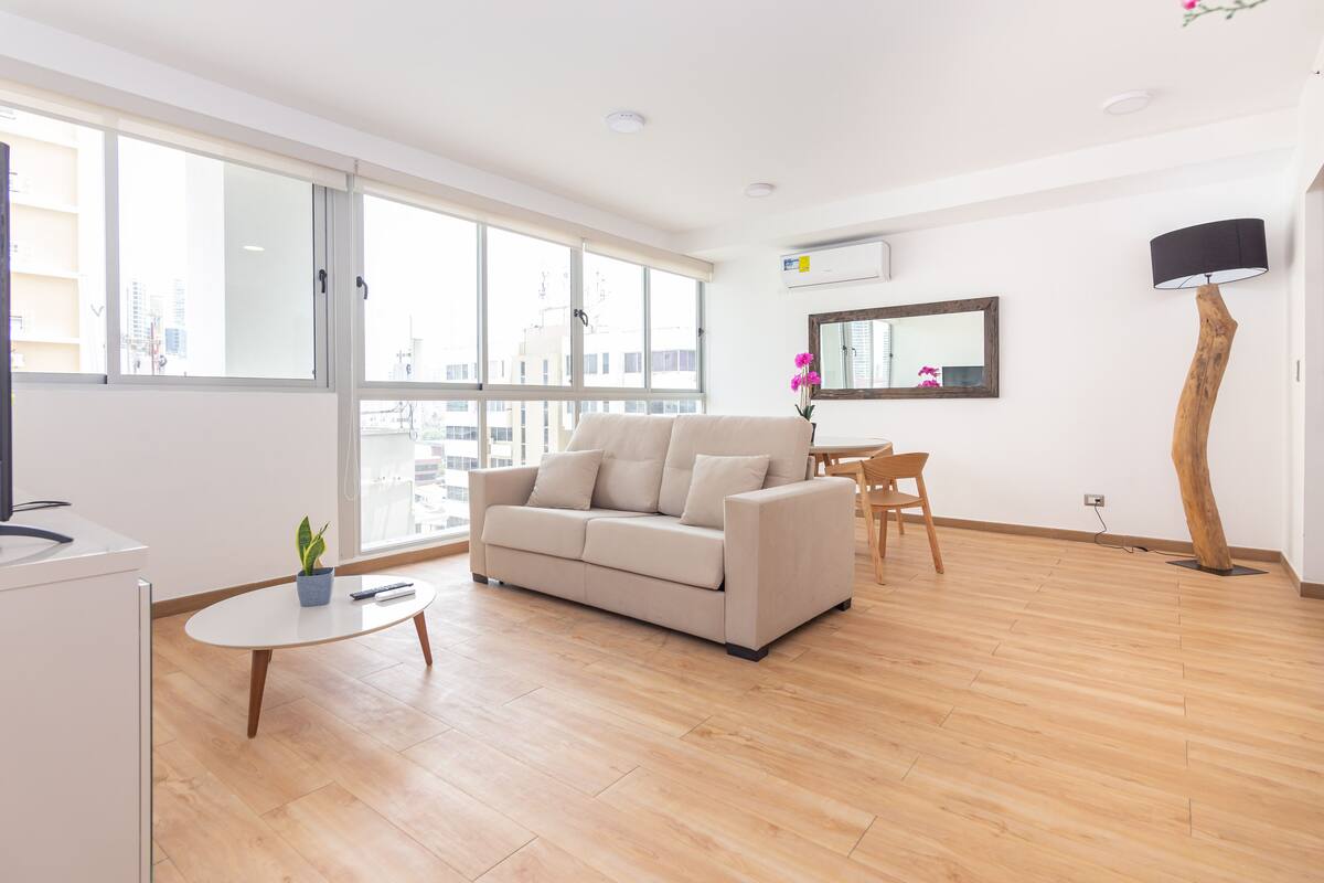 A modern living room with a sofa, a small table, and a chair. There is a large window providing a view of buildings outside.