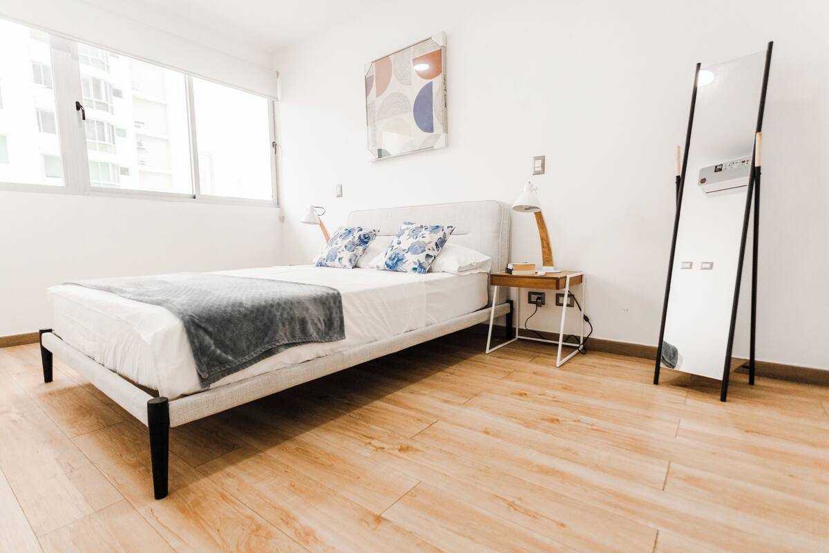 A modern bedroom with a bed, a nightstand, a lamp, and a large window.