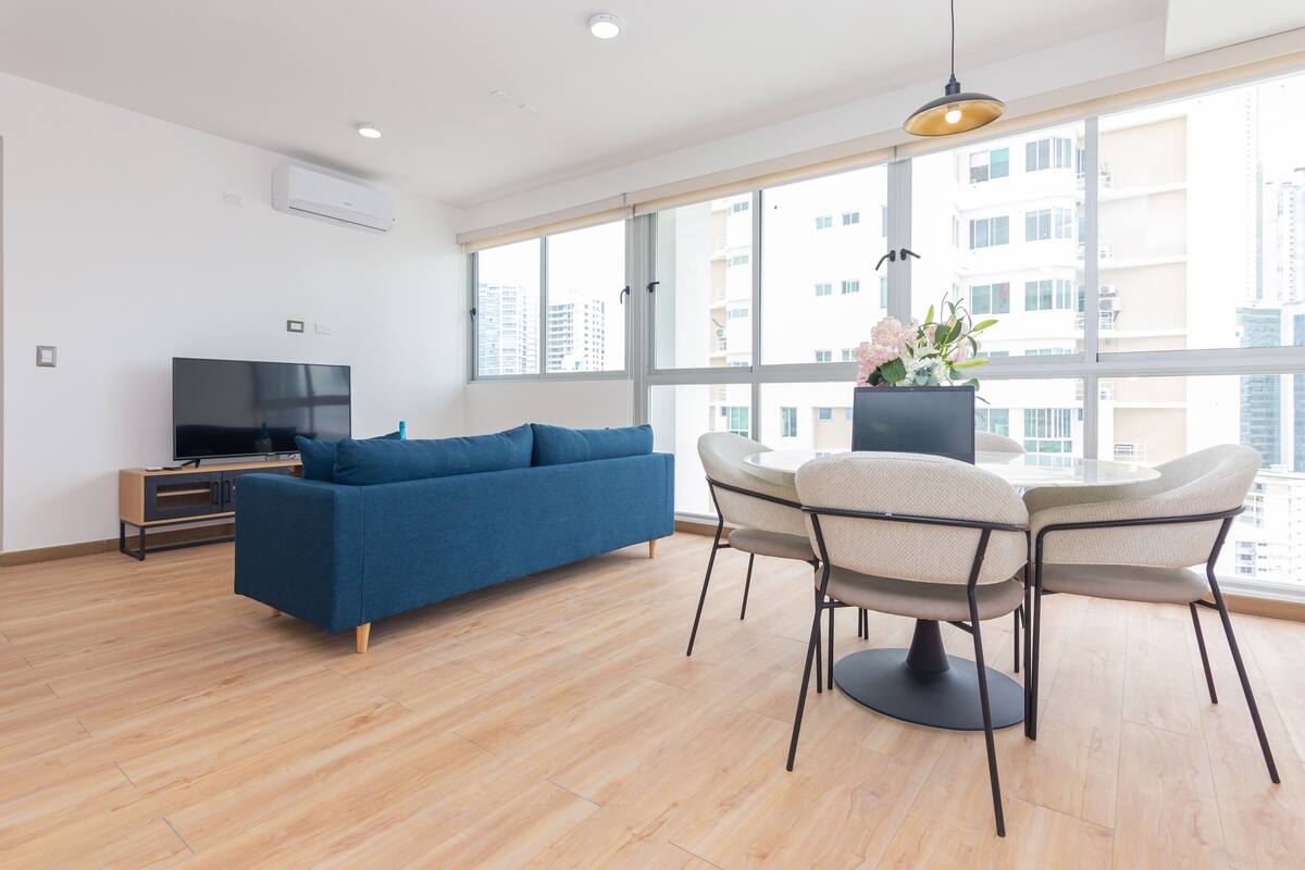 A modern living room with a blue sofa, a dining table with chairs, a TV on a stand, and large windows offering a city view.