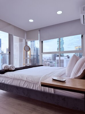 Een moderne hotelkamer met een groot raam, een bed met witte lakens, een houten nachtkastje en uitzicht over de stad.