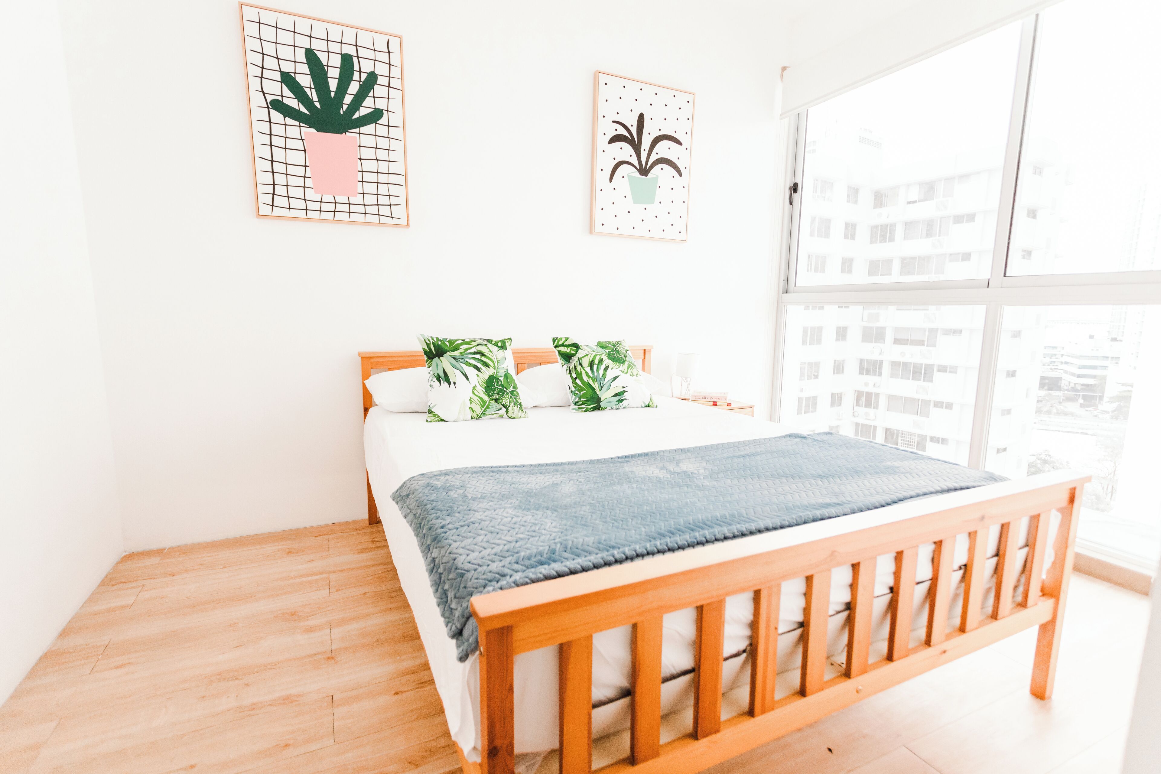 A bedroom with a wooden bed, white bedding, and green and white patterned pillows. There are two framed plant illustrations on the wall.