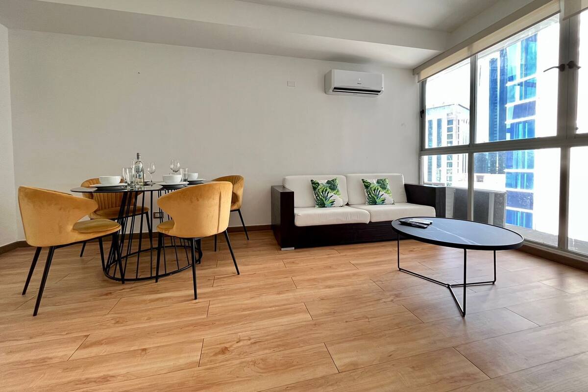 A modern living room with a dining area, large windows, and a sofa with tropical cushions.