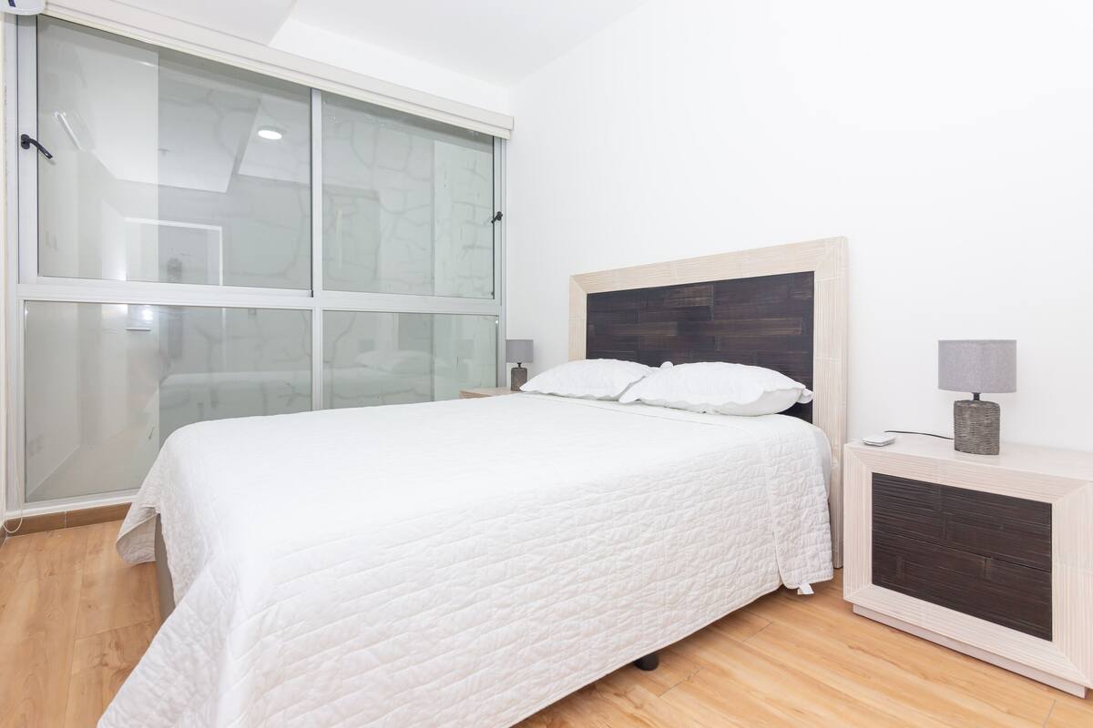 A bedroom with a large window, a bed with white bedding, a bedside table with a lamp, and a small heater.