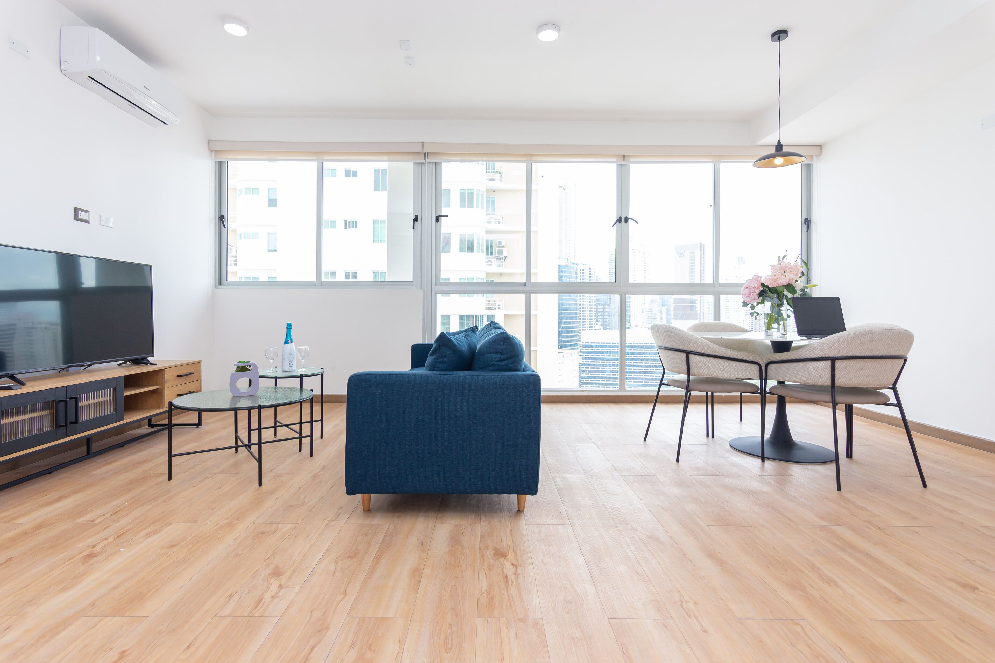 Una sala de estar moderna con un sofá azul, una mesa de comedor con sillas, un televisor y un ventanal con vistas a la ciudad.