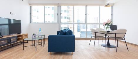 Una sala de estar moderna con un sofá azul, una mesa de comedor con sillas, un televisor y un ventanal con vistas a la ciudad.