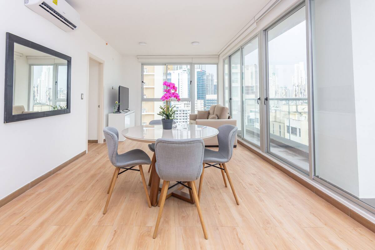 A modern dining area with a round table and four chairs, a potted orchid, and a large window with a city view.