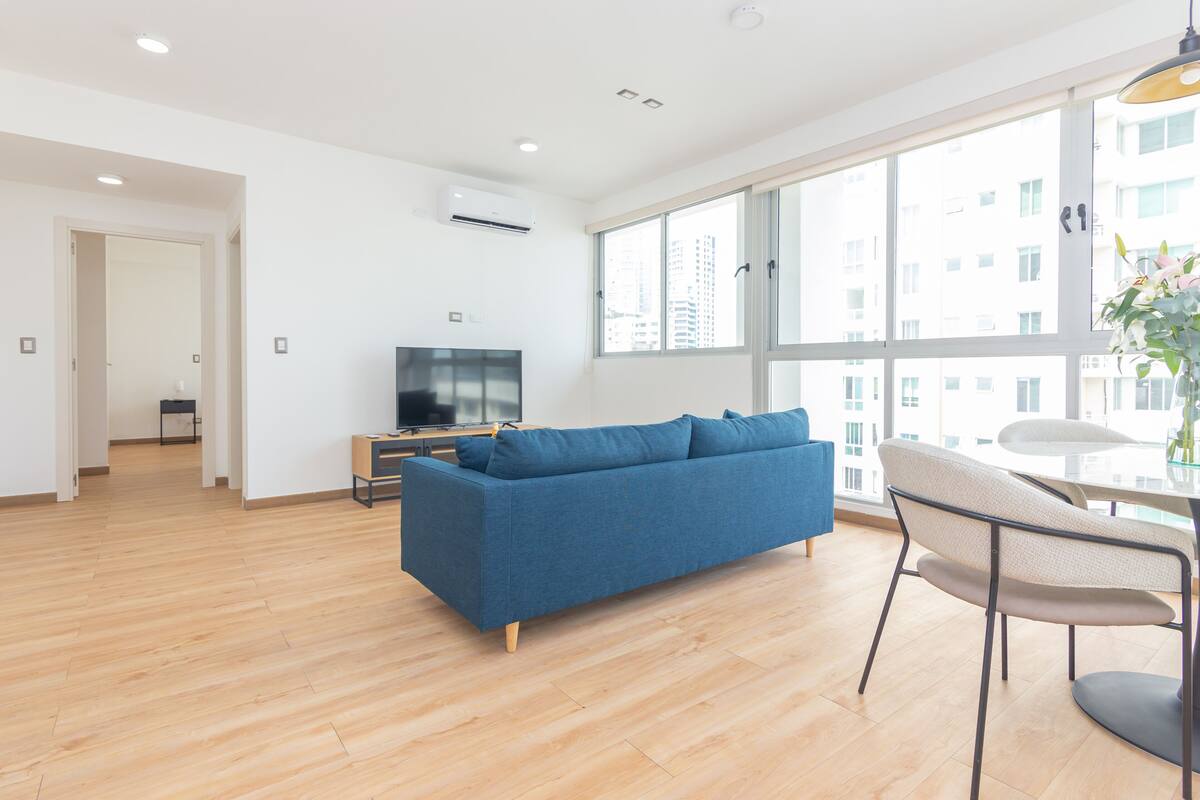 A modern living room with a blue sofa, wooden flooring, a TV, and large windows offering a city view.