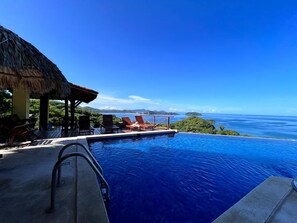 Une piscine à débordement offrant une vue sur l’océan et une structure à toit de chaume dotée de chaises longues.