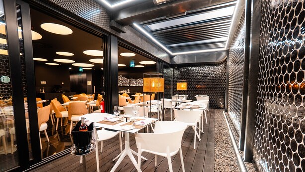 A modern restaurant interior with round tables and chairs, a patterned wall, and a ceiling with linear lighting fixtures. - Hills Hotel Lisboa (Lisbon)