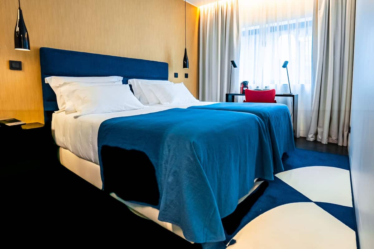 A hotel room with a large bed, two bedside tables, a red lamp, and a window with curtains.