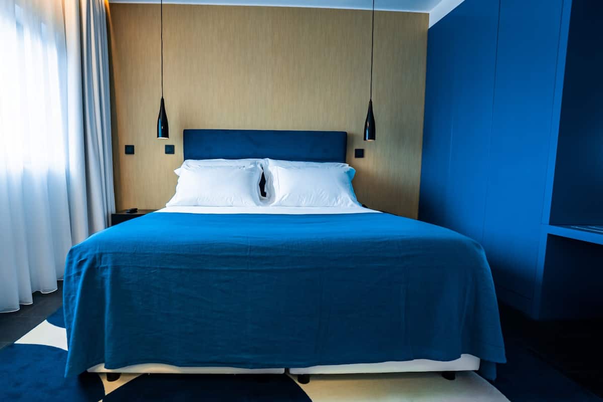 A hotel room with a large bed, a wooden headboard, two pendant lights, and a view of the outdoors through sheer curtains.