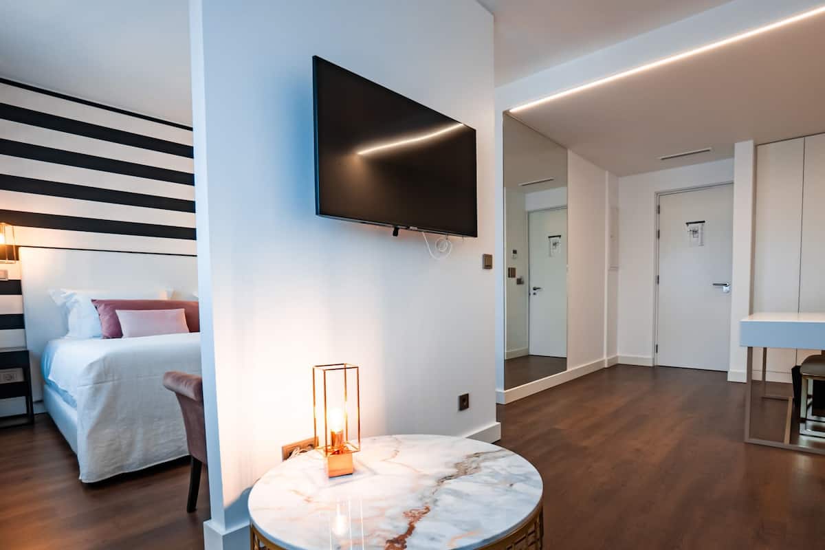 A modern hotel room with a bed, a round marble table, a TV mounted on the wall, and a hallway leading to a bathroom.