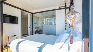 A modern bedroom with a large bed, a glass partition, a bathroom with a sink and mirror, and a decorative hanging light fixture. - Hills Hotel Lisboa (Lisbon)