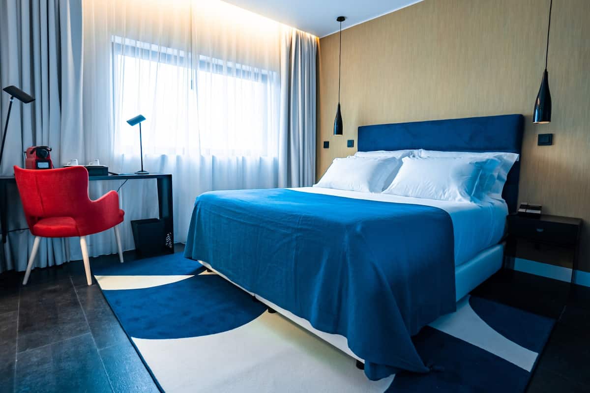 A modern hotel room with a large bed, a desk, a red chair, and a desk lamp.