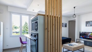 Ett modernt kök med en inbyggd ugn, en matplats med ett träbord och en lila stol, och ett vardagsrum med en TV och en soffa.