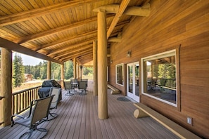 A covered wooden deck with outdoor furniture, large windows, and a view of the surrounding forest.