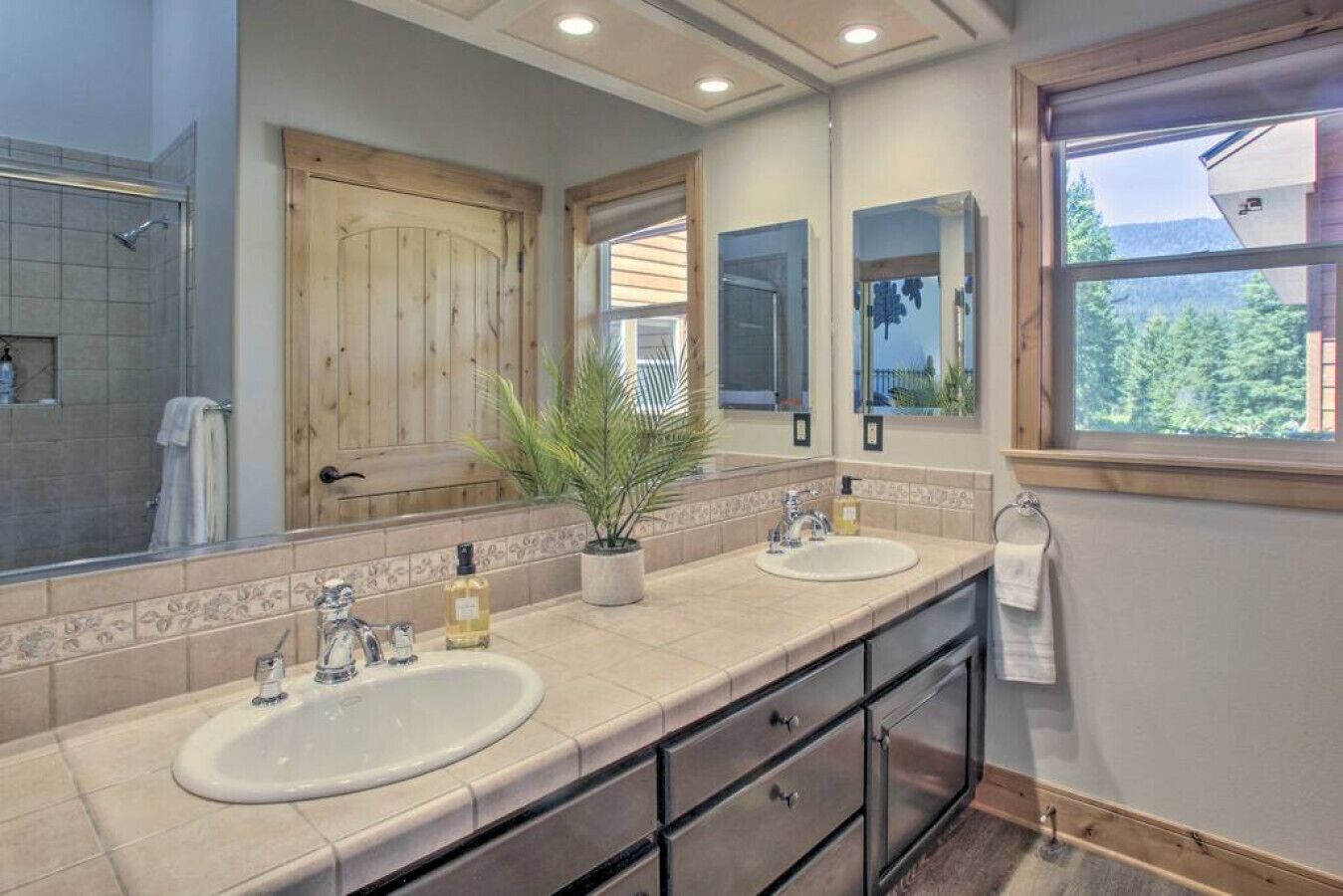 Une salle de bain avec deux lavabos, un grand miroir et une fenêtre donnant sur les arbres.