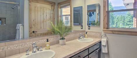 Une salle de bain avec deux lavabos, un grand miroir et une fenêtre donnant sur les arbres.
