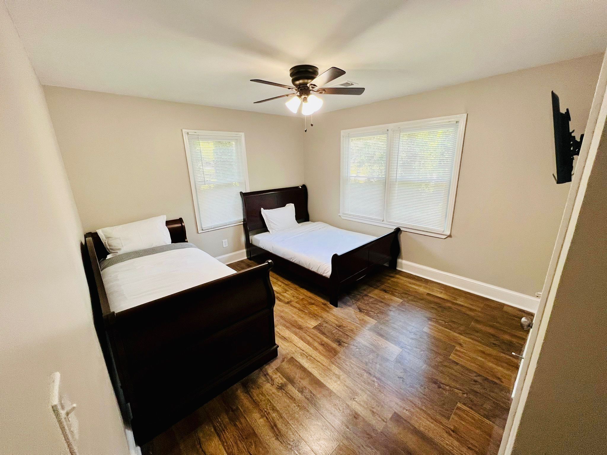 A bedroom with two beds, wooden flooring, a ceiling fan, and windows with blinds.