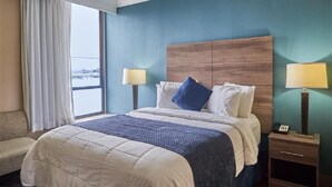 A hotel room with a large bed, a wooden headboard, two bedside tables with lamps, a window with curtains, and a view of the city. - Queen Bed Standard View (Fairhaven)