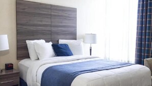 A hotel room with a large bed, a wooden headboard, two bedside lamps, and a window with curtains. - Queen Bed Standard View (Fairhaven)