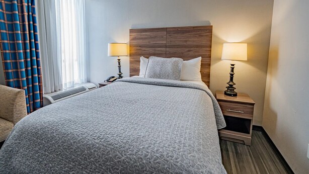 Habitación de hotel con una cama grande, dos mesitas de noche con lámparas, un cabecero de madera, una silla y una ventana con cortinas.