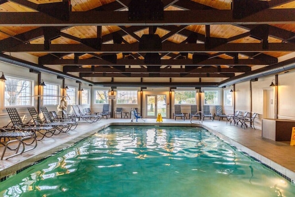 An indoor swimming pool with wooden beams, lounge chairs, and large windows.