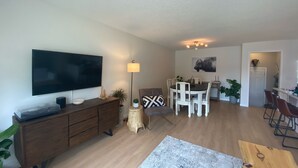 Un salon moderne doté d’un téléviseur à écran plat, de meubles en bois, d’un coin repas avec des chaises blanches et d’un petit espace cuisine.