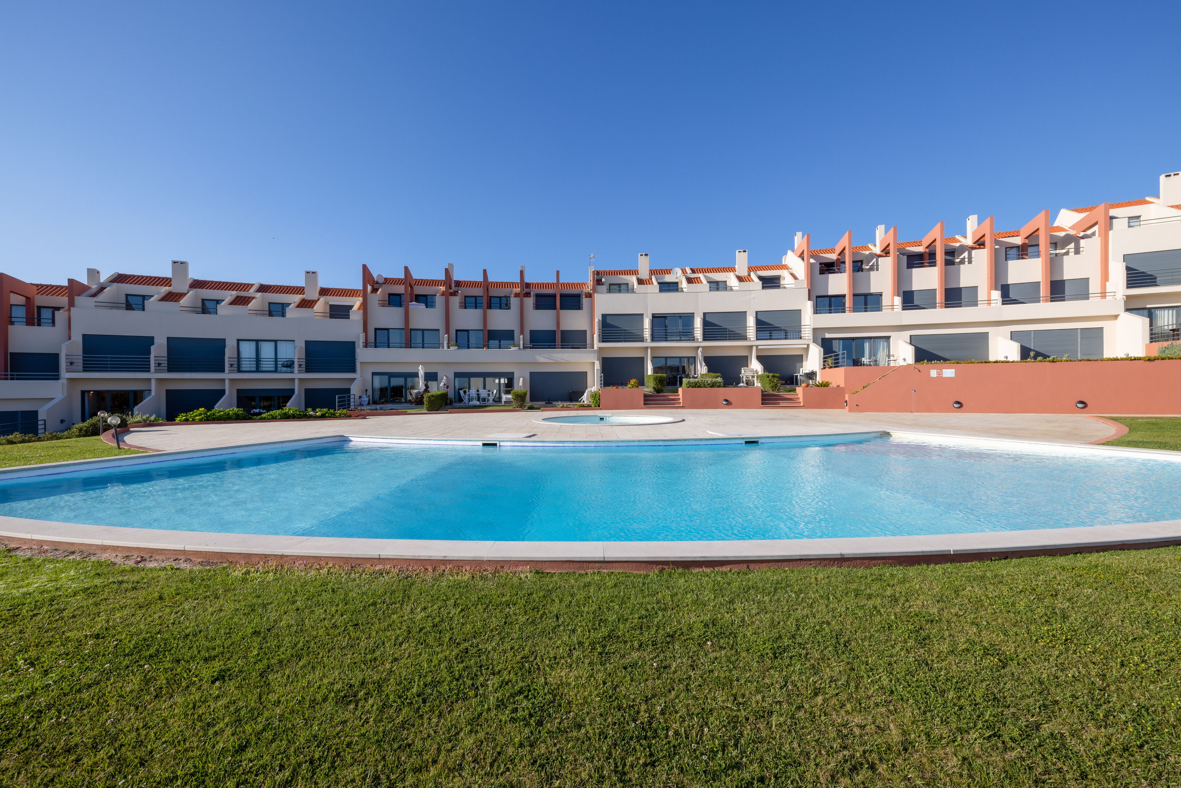 A modern hotel with a large swimming pool, multiple balconies, and a well-maintained lawn.