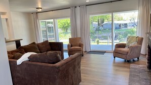 A spacious living room with a brown sofa set, a smaller armchair, and a fireplace. Large windows offer a view of the outdoors.