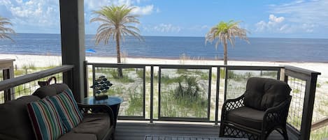 Una terraza cubierta con una mesa de madera y dos sillas, con vistas a una playa con palmeras y el océano.