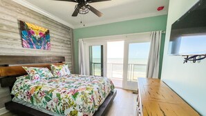 A bedroom with a bed, a wooden headboard, a ceiling fan, a colorful painting, and a view of the beach through a sliding glass door. - Beachfront--Beachfront- Driftwood Yard (Mexico Beach)