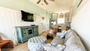 A living room with a grey sofa, ottoman, and wicker chair. There is a television on a cabinet, a kitchen in the background, and a dining area with a table and chairs. - Beachfront--Beachfront- Driftwood Yard (Mexico Beach)