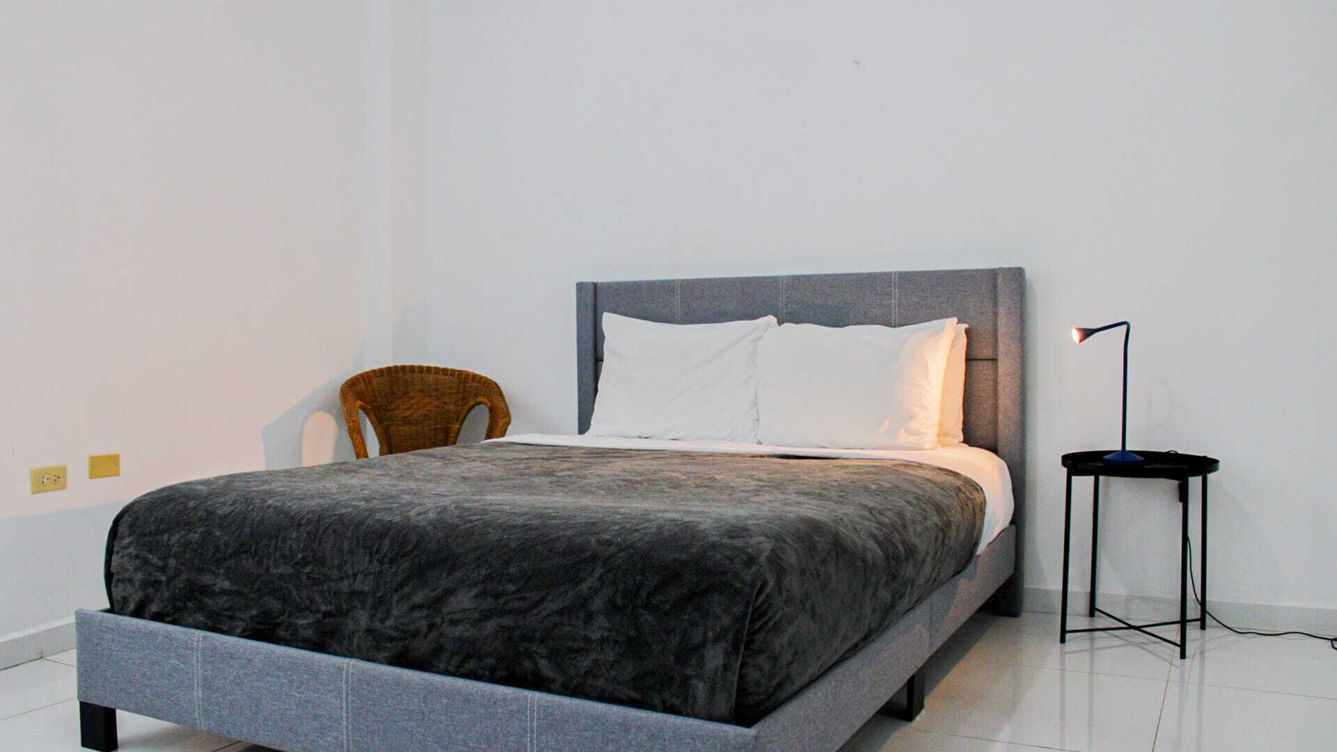 Ein modernes Schlafzimmer mit einem grauen Bett, weißen Kissen, einem dunkelgrauen Kopfteil, einem Korbstuhl und einer Stehlampe.