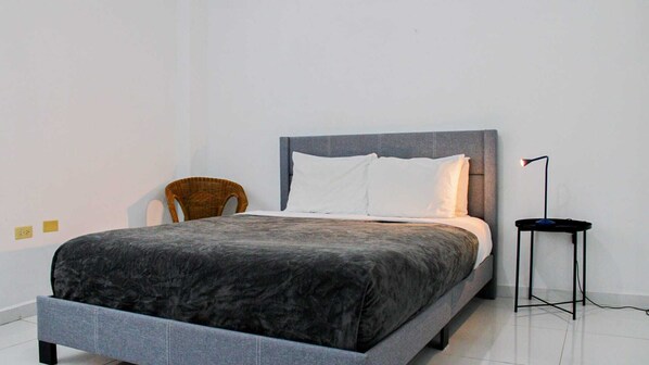 A modern bedroom with a grey bed, white pillows, a dark grey headboard, a wicker chair, and a floor lamp. - Arecibo Hotel 2104 (Arecibo)