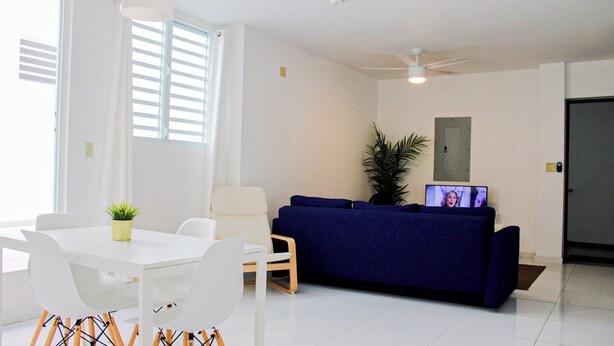 A modern living room with a white dining table, white chairs, a blue sofa, and a ceiling fan. - Arecibo Hotel 2104 (Arecibo)