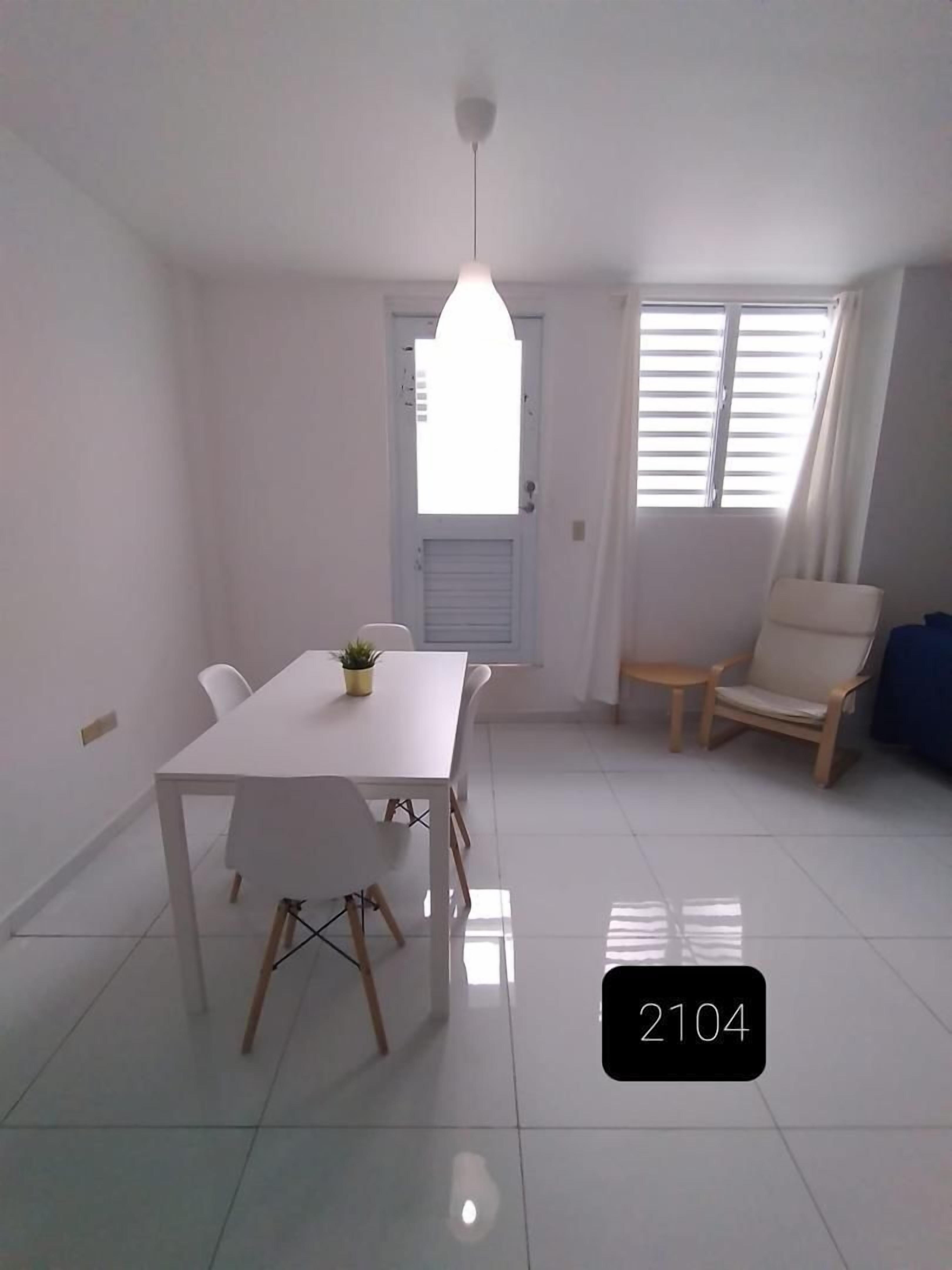 Una sala de estar moderna y bien iluminada con comedor, que incluye una mesa blanca, sillas y una lámpara colgante.